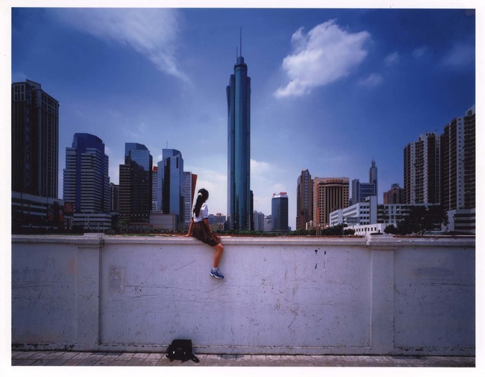 A long-haired youth sits on a wall with their back to the camera, looking out towards downtown Shenzhen.