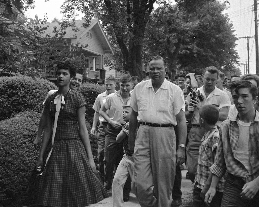 A young black woman in a checkered dress is followed by a group of white men
