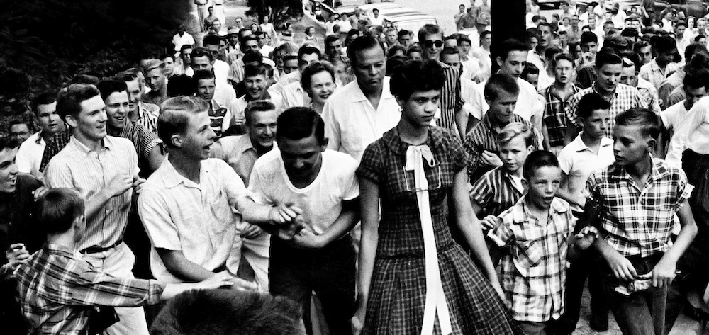 A young black woman in a checkered dress is surrounded by a white and mostly male mob