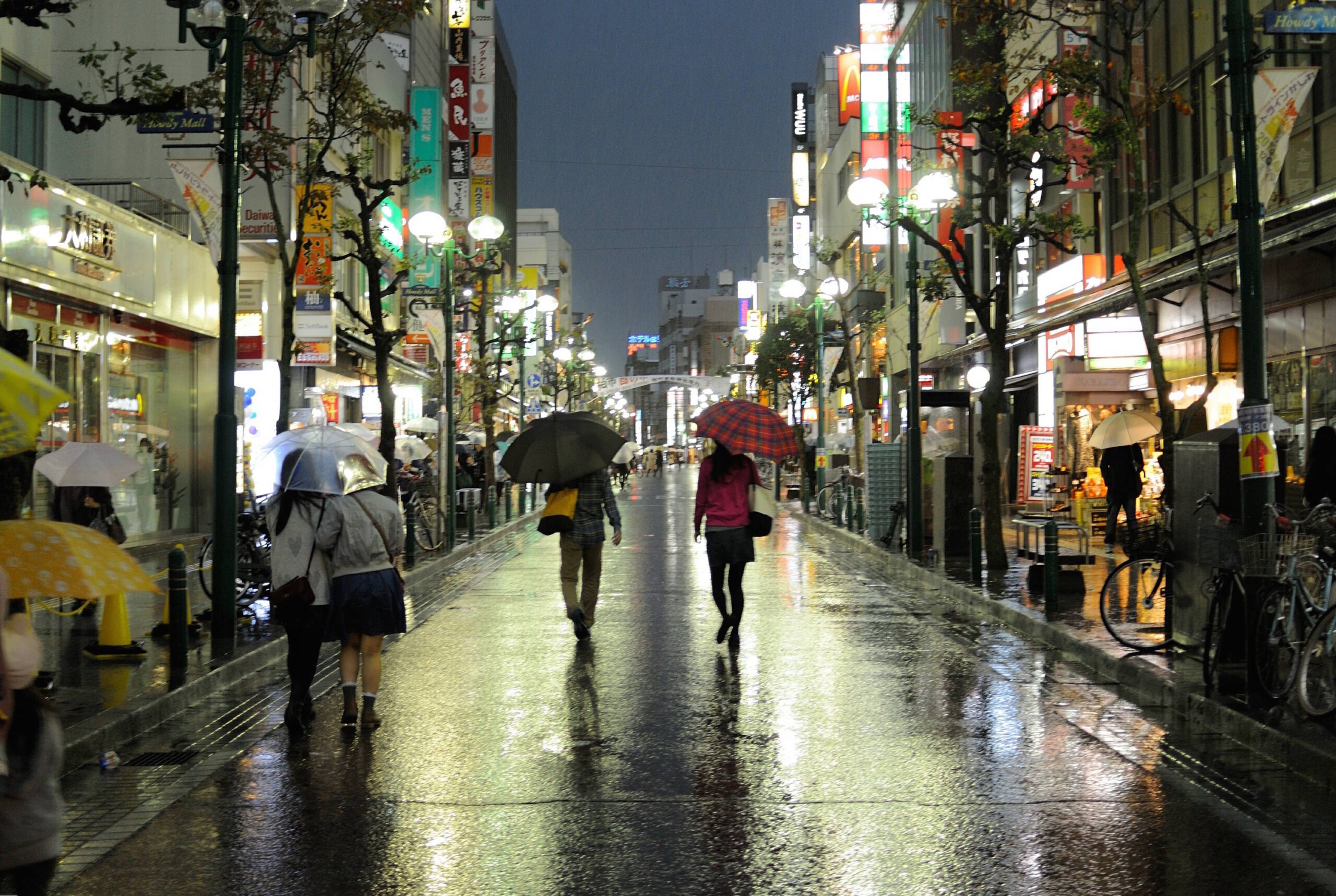 Pedestrians hold umbrellas, their backs to us, walking on a Tokyo street on a rainy night.