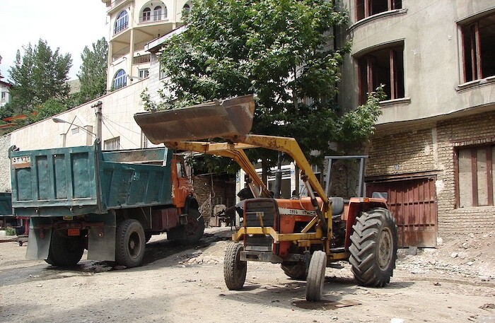 A green dump truck and a yellow and orange tractor in front of an apartment building on a dirt road.